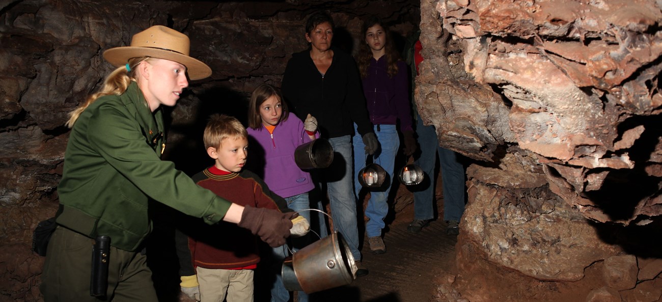 A ranger with a family in a dim cave passage, all are holding candle buckets.
