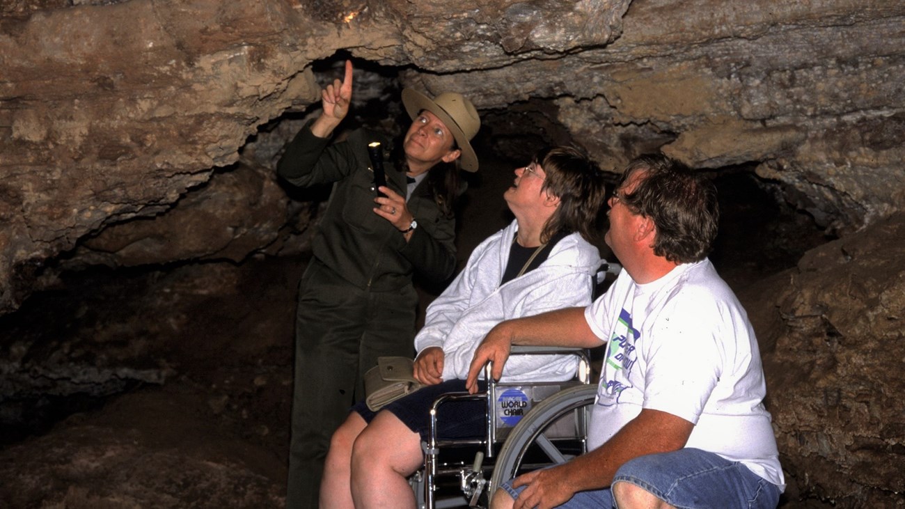 a ranger with two visitors in a cave room looking at a formation, one visitor is in a wheelchair