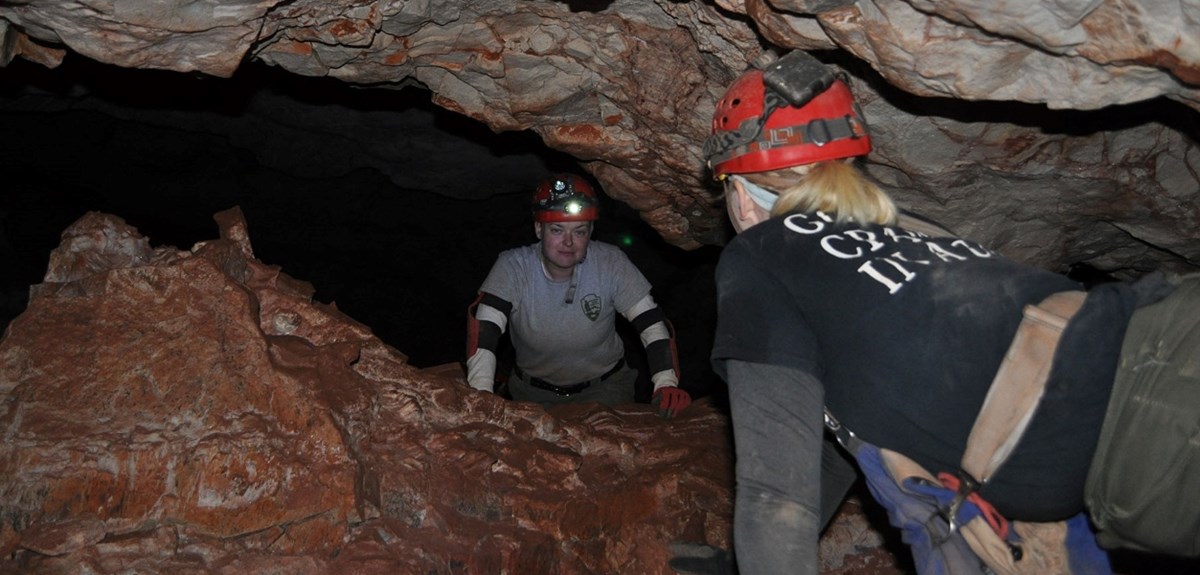 two people with helmets, headlamps, and caving clothes crawl through a low cave passage