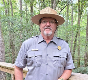 Steve Akins A Caucasian appearing man in a uniform leans on a railing and smiles for the camera. A forest is in the background.