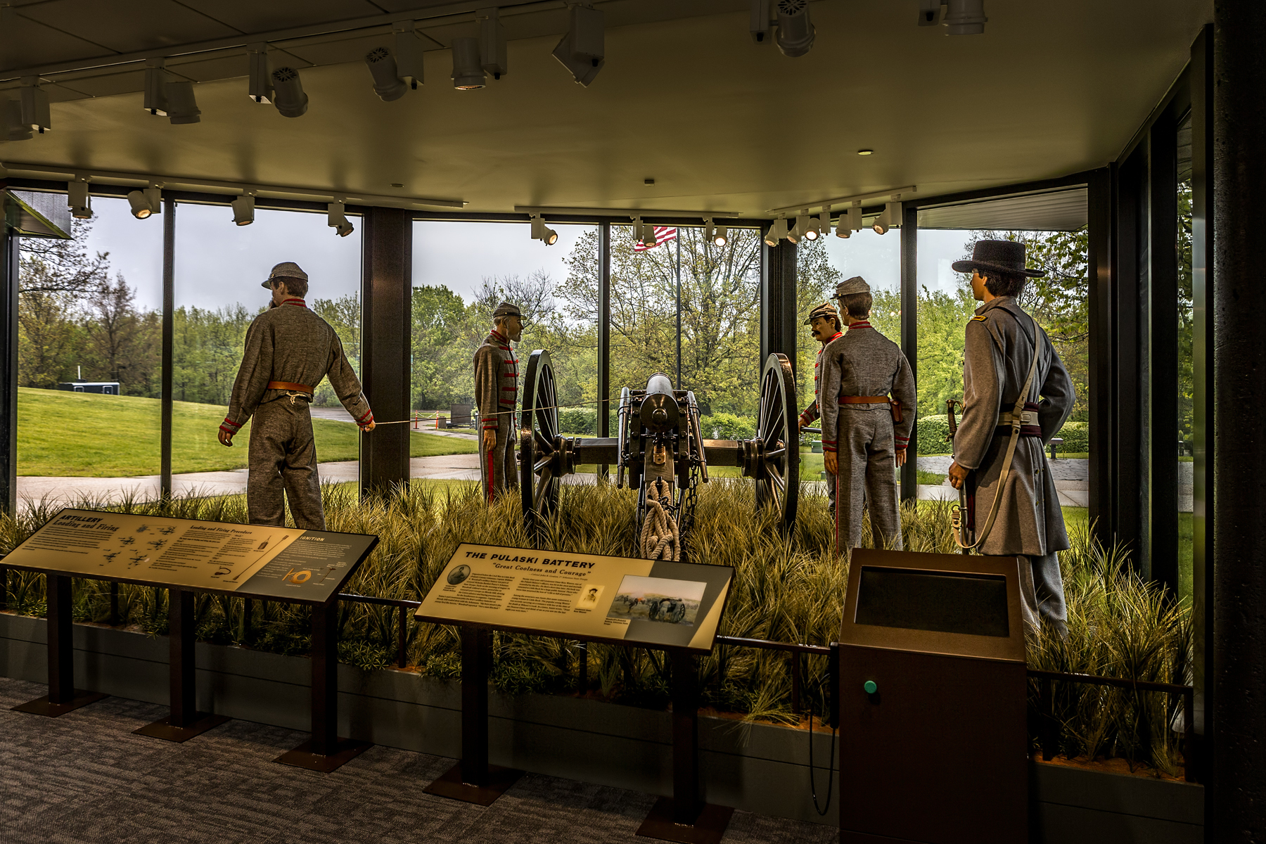 The Pulaski Battery Visitor Center window display featuring a grassy field with 5 cannoneer mannequins in period dress representing The Pulaski Battery in the position of fire at the cannon.