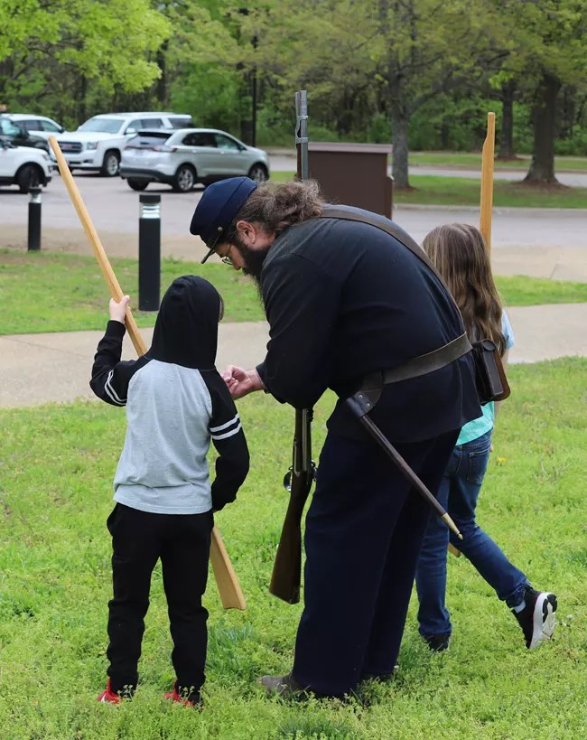 Man wearing a civil war uniform helps child in a hoodie hold a wooden musket.