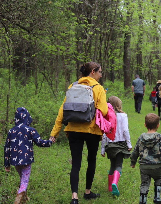 A mother holds the hands of two children in coats on a hike, facing away from the camera.