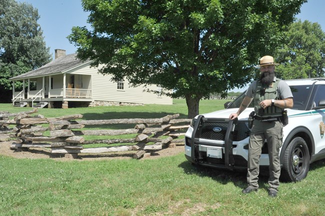 Law enforcement officer stands in front of his squad car. The Ray House and split rail fencing are in the background.