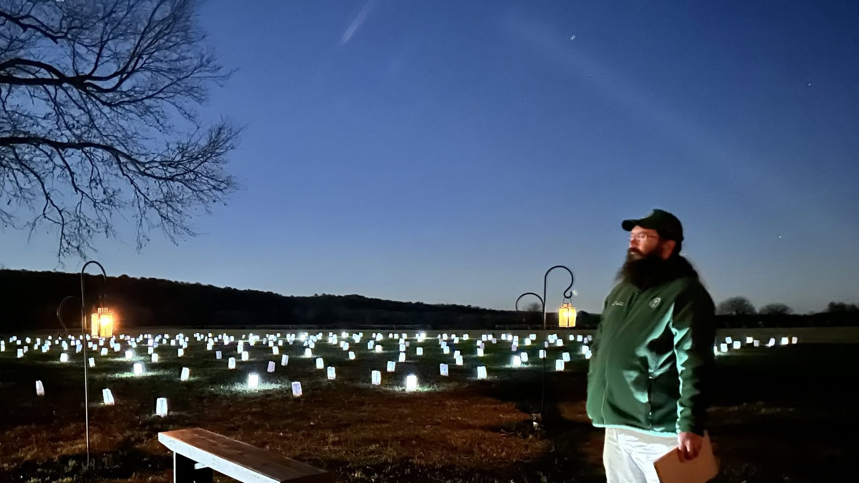 A volunteer with a bushy beard and glasses stands in front of a field full of luminary lanterns near dusk as the last light of dusk lights up clouds and outlines a tree.