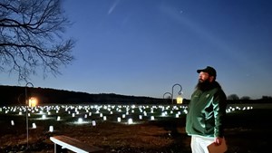A volunteer with a bushy beard and glasses stands in front of a field full of luminary lanterns near dusk as the last light of dusk lights up clouds and outlines a tree.