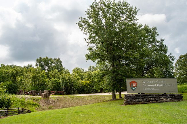 Entrance gate of the park. Park sign reading Wilson's Creek national battlefield with NPS logo sits infront of a large tree.
