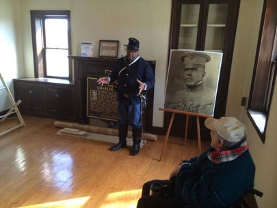 A man wearing a blue army uniform and a blue cap, speaks to people as he stands on a wooden floor next to a portrait of a soldier.