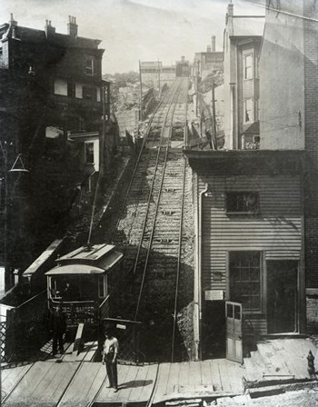 B&W photo of a trolley car and a rail line ascending up a hill between buildings