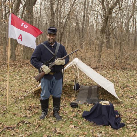 A man standing next to a troop flag and in front of a small tent holding a Sharps carbine