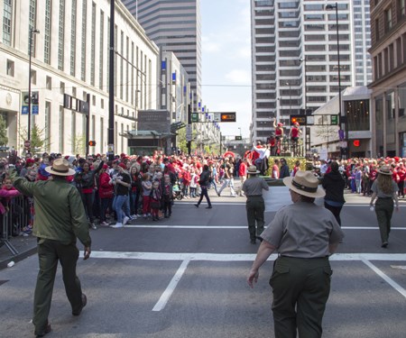 Several rangers in green pants, grey shirts and flat hats walk between crowds of people on sidewalks Several rangers in green pants, grey shirts and flat hats walk between crowds of people on sidewalks