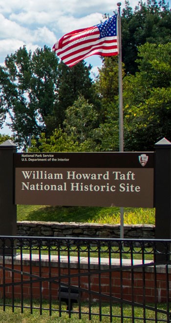 An American Flag on a flagpole being blown by the wind from right to left, behind a sign reading William Howard Taft National Historic Site