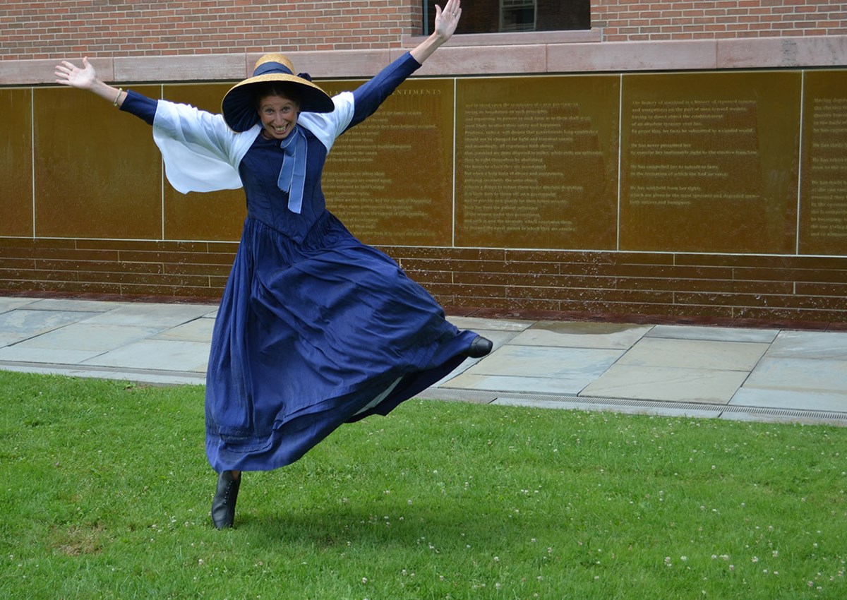 An excited young woman stands on one foot, with arms stretched wide. She poses in front of the Declaration of Sentiments waterwall in a historical-style blue dress and bonnet.