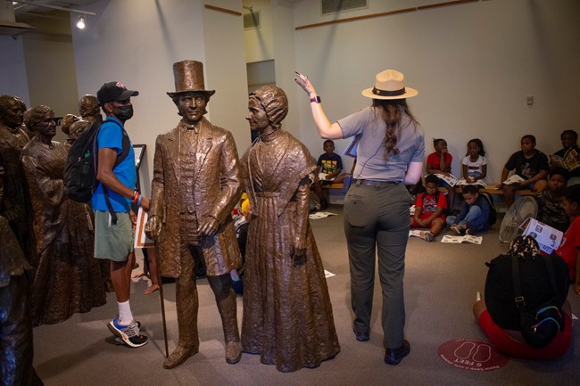 A Ranger stands next to a bronze statue of a man and woman, and talks to a group of kids.