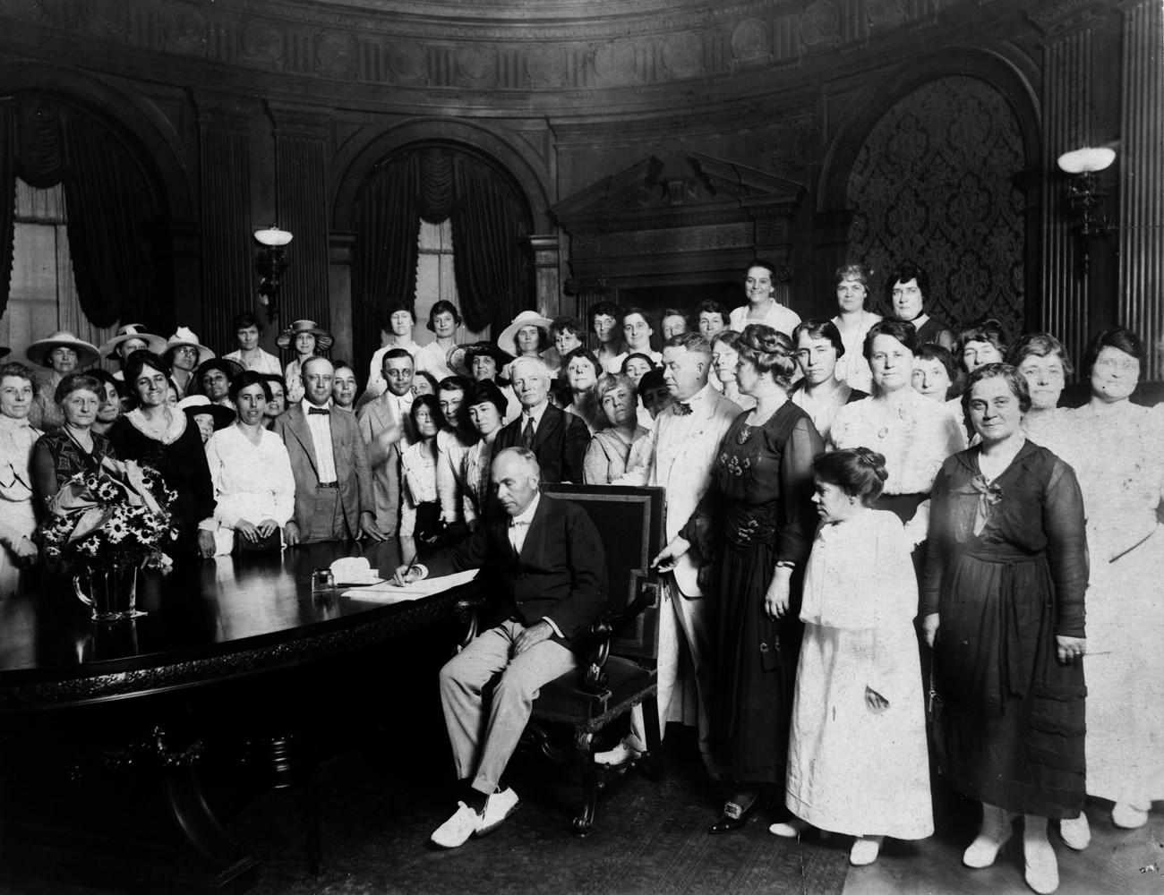 A historical photograph of a man in a crowded room surrounded by people signing a document.
