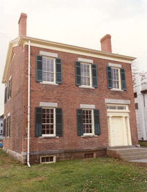 The M'Clintock House is a red brick residence with three equally spaced windows facing front on the second story, with two windows and the front door on the first story. Green shutters frame each window and four stone stairs mark the entry to the pail yellow front door on the right.