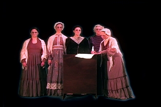 Five women, dressed in 1800's clothing, are gathered around a central figure reading.