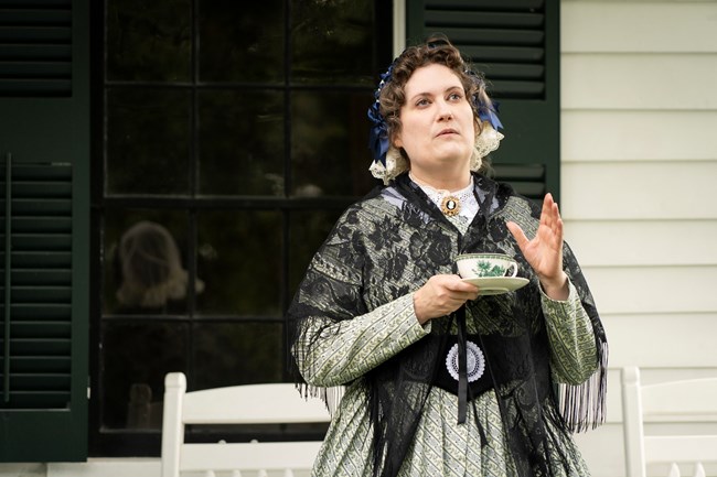 A woman in 19th century clothes stands holding a teacup on the porch of a house.