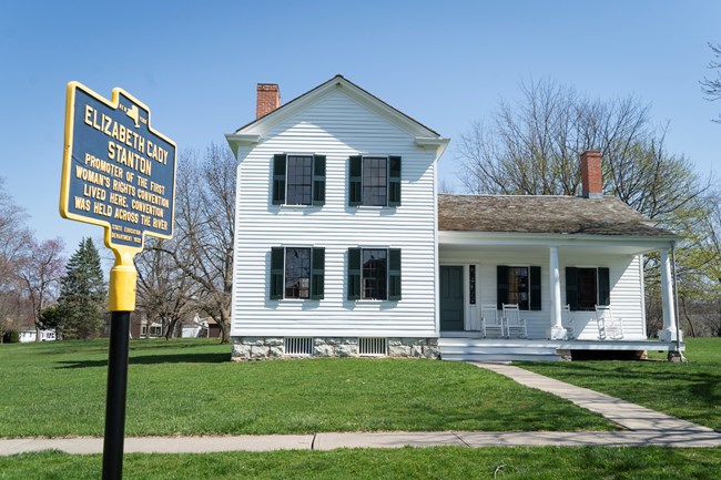 A two-story white house with a yellow and blue historic marker out front.