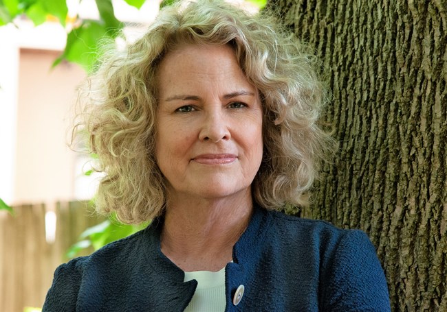 Dr. Kate Clifford Larsen portrait. She has blonde, curly hair, and is wearing a white shirt under a blue coat. She leans on a tree trunk.