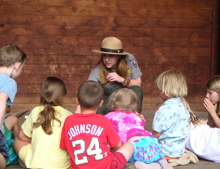 A park ranger crouches down to talk to a seated group of small childern