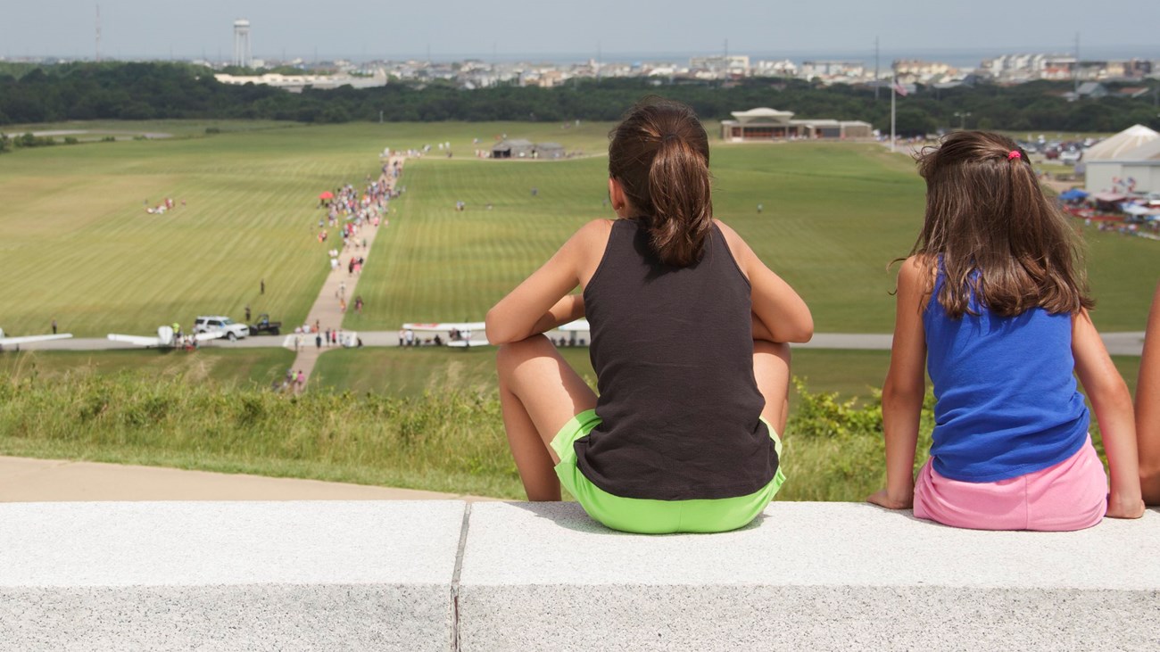 Two children sit overlooking field with small buildings.