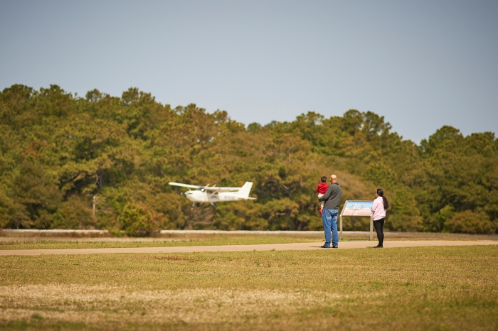 Two adults and one young visitor watch a white airplane land.