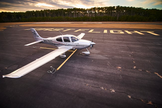 Small propeller plane parked beside a runway with the words "First Flight" written on the asphalt