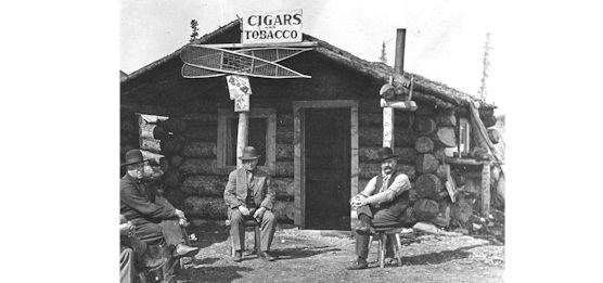 Historic photo of men sitting on stools in front of log cabin store.