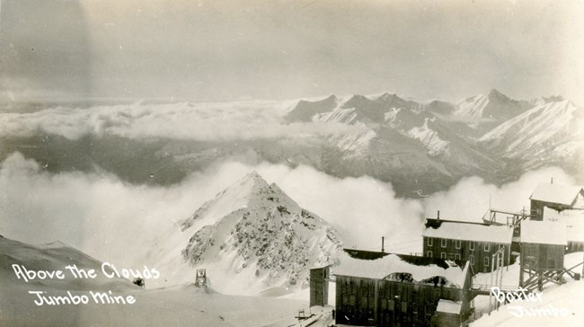 Black and white sepia vintage photo of Kennecott town with clouds below over the glacier and words on the image, Jumbo Mine Above the Clouds.