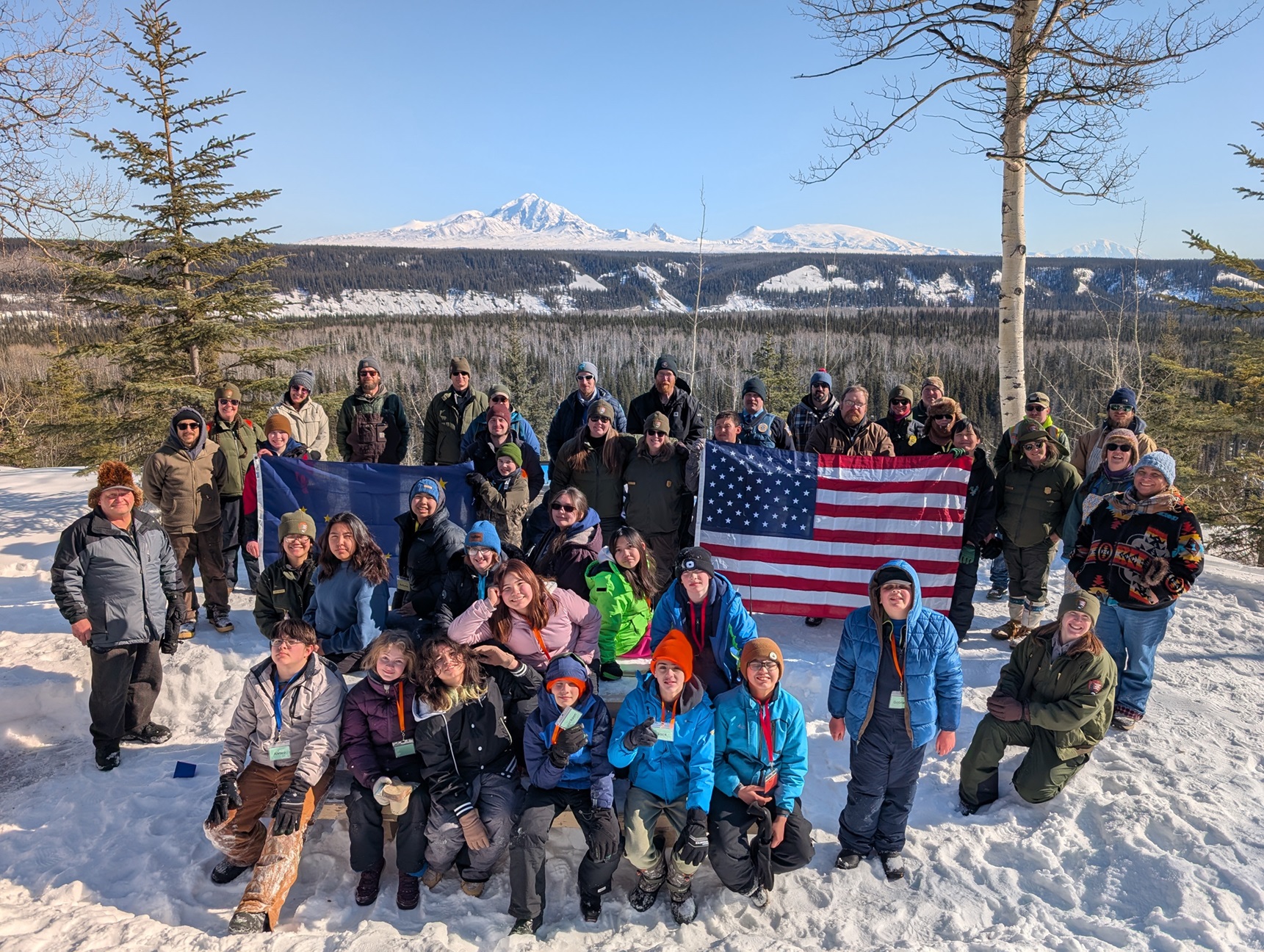 Copper River School District Students, National Park Service staff, and partners at Copper Center bluff overlook with the Wrangell Mountains in the background on a sunny day.