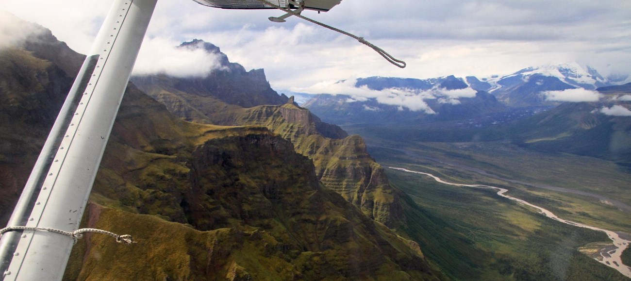 View from a bush plane window overlooking the Black Hills, a braided river, and the snow capped mountains beyond.