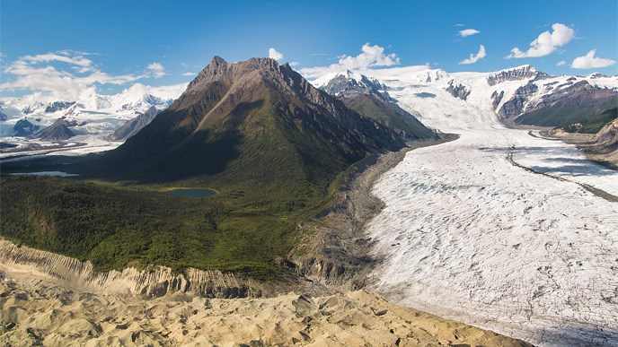 View of glaciers with forest, mountains, and cloudy blue skies in background.