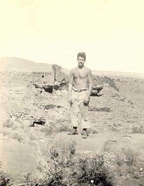 A black and white photo of a man, bare chested, stands in front of a desert landscape and a pueblo.