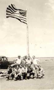 A black and white photo with a group of 11 men in two rows posting in front of a flying American Flag with a barren landscape and the front of an old truck in the background.