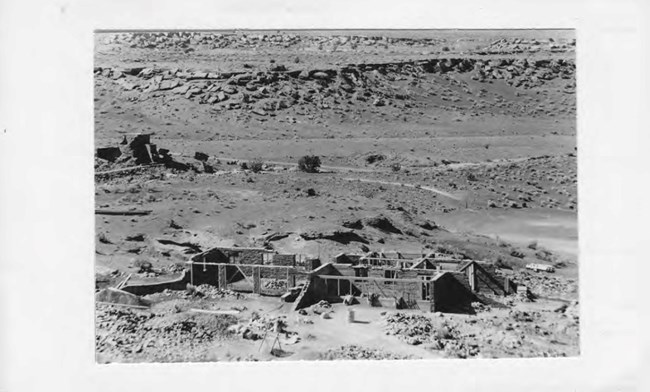 A black and white photo from the 1930s of a stone house under construction with a pueblo in the background.