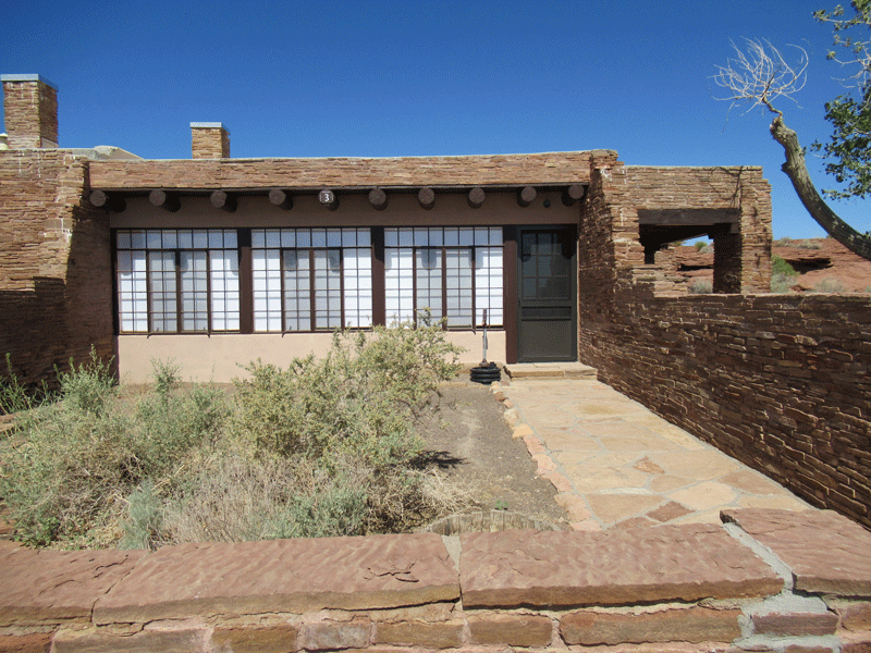 The exterior entrance of a sandstone Pueblo Revival style building with three large paned windows and a small courtyard.