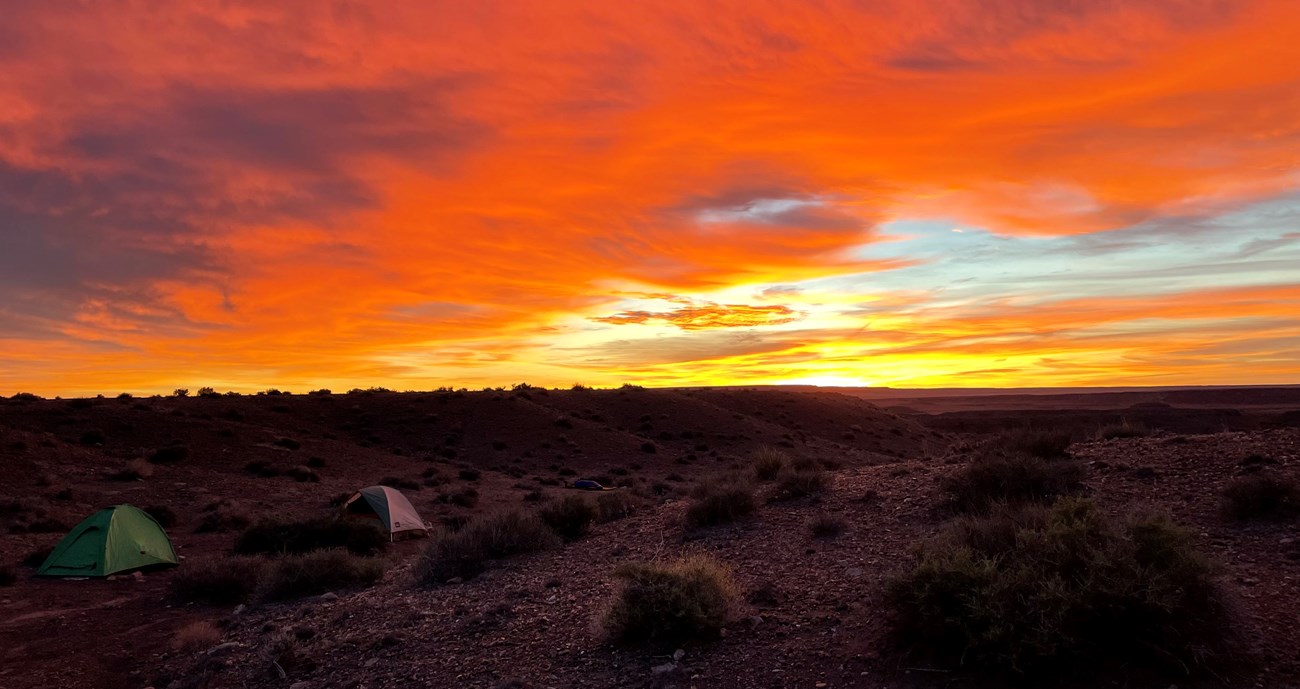 Two domed camping tents set up among black cinders and red sandstone with a bright yellow and red sunrise in the background.