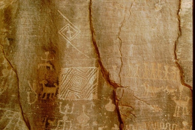 An old photograph of geometric, zoomorphic, and anthropomorphic designs pecked into the surface of a vertical, orange rock face. The rock is also criss-crossed by several diagonal cracks.