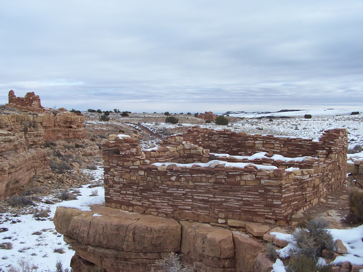 Three partial pueblo structures made out of limestone next to a small box canyon. There is snow on the ground.