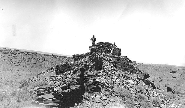 Two men standing on the top of Wupatki pueblo in the early 1900s before archeologists did any reconstruction or excavation. Only partial walls existed and the remaining stones were on the ground below.