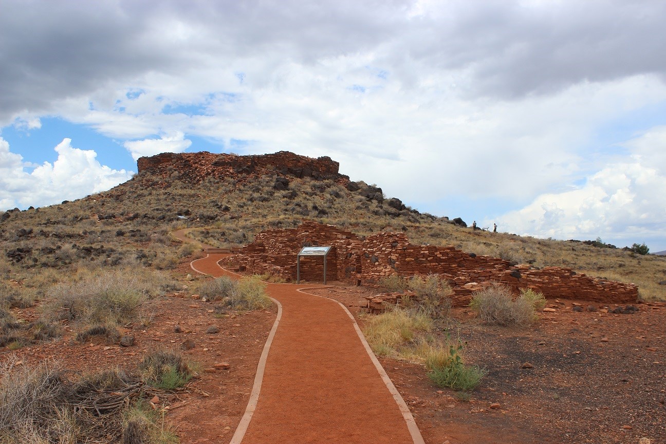 An accessible path leading past one small stone pueblo on the right with a larger pueblo at the top of a small hill in the background.