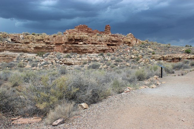 A box canyon pueblo on a cliff above with a rough gravel trail below.
