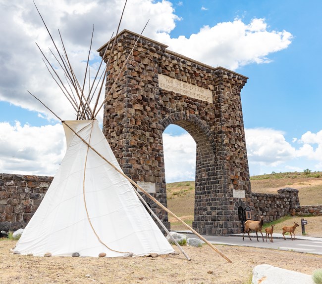 three elk walk through a large, stone archway with a large teepee nearby