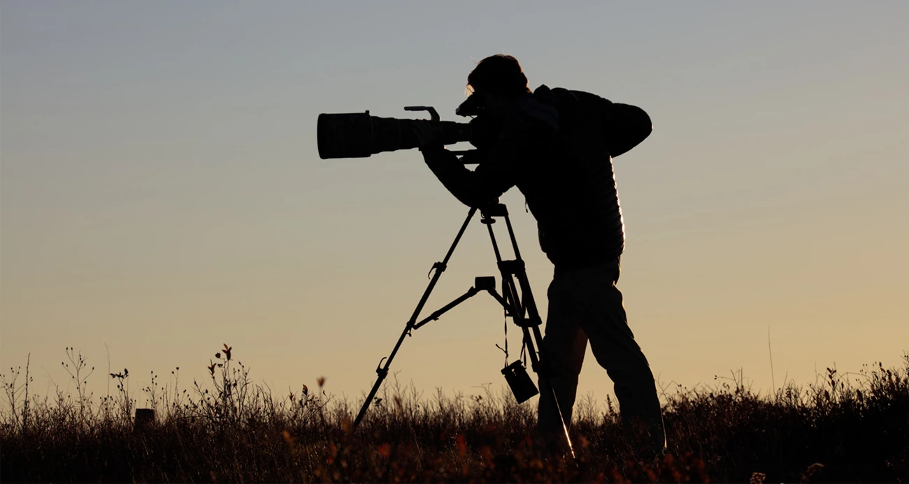 A photographer looks into their camera with a telephoto lens, while silhouetted against the sun
