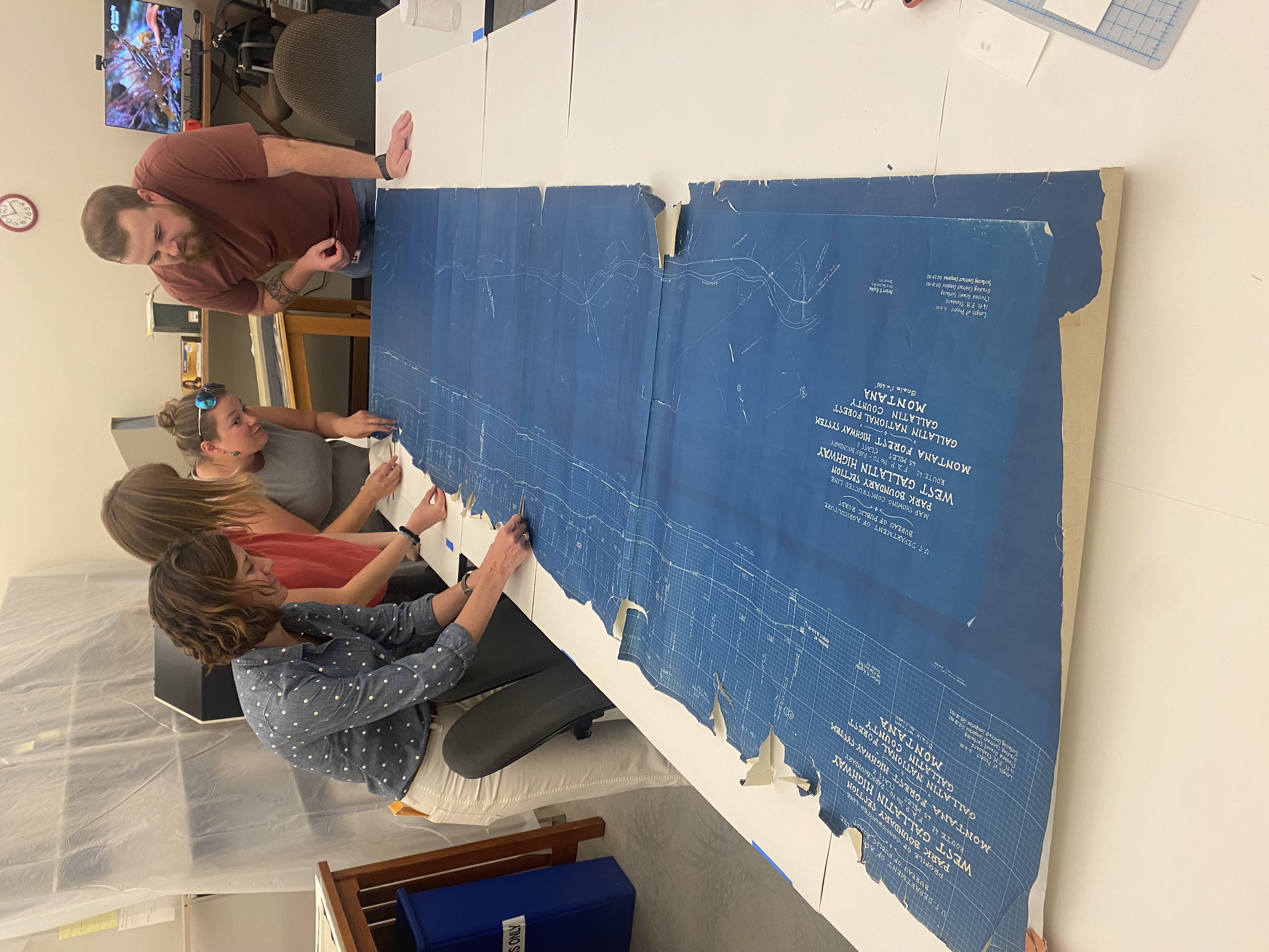 Four staffmembers lean over a long blueprint to mend the torn edges