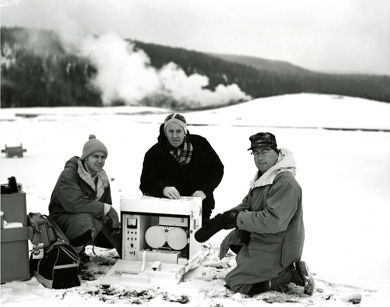 An historic black-and-white photograph showing three researchers by an instrument in the snow