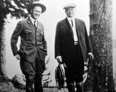 A man in an early park ranger uniform next to a man holding fish and a fishing rod