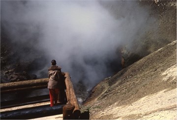 A person stands in front of rising steam from a large vent in a hillside above blue water