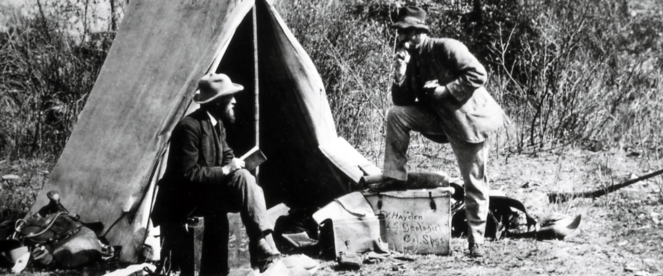 A man rests his elbow on a raised leg next to a man sitting in front of a primitive tent A man rests his elbow on a raised leg next to a man sitting in front of a primitive tent